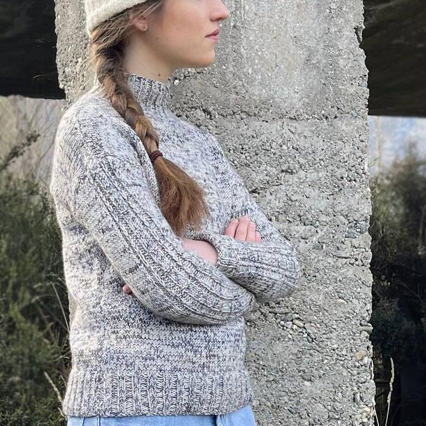 Young women leaning against concrete pole wearing a hand knitted marle sweater with sleeve details and cream beanie