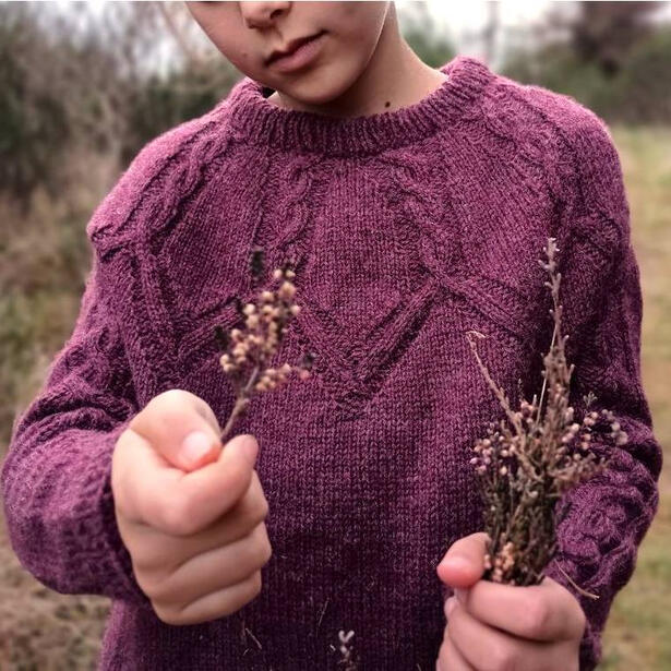 Girl wearing a hand knitted cable yoke seater of muted rose red colour and holding a small bouquet of wild grassland flowers