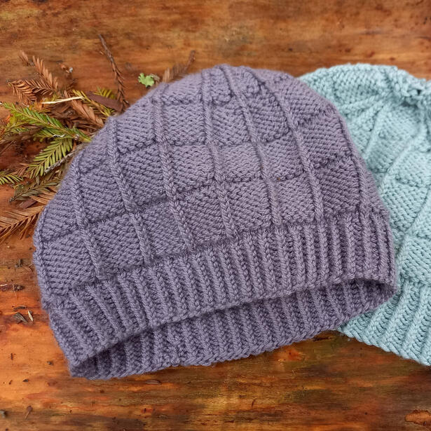 Purple knitted beanie with square pattern on wooden background with another light blue beanie behind