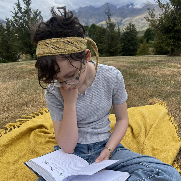 Young girl wearing a hand knitted yellow headband with diagonal lacework sitting on a blanket in a field near mountains